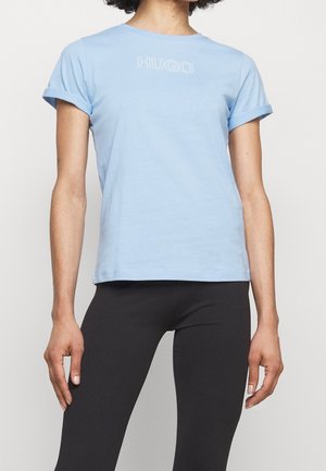 Osoba nosząca jasnoniebieski T-shirt z krótkim rękawem z nadrukiem "HUGO" na piersi oraz czarne dopasowane spodnie na jednolitym tle.