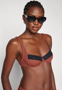 Rjav bikini vrh z črnimi detajli, strukturiran dizajn, debele naramnice in teksturirana tkanina. Model nosi črne mačje očala.