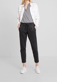 Witte denim jack over een zwart-witte geruite blouse, gecombineerd met tapered zwarte broek en witte sneakers. Opvallende tailleband met trekkoord.