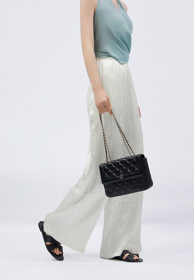 Borsa a tracolla in pelle nera con motivo trapuntato, cinturino a catena e logo; indossata con una maglietta azzurro chiaro e pantaloni larghi bianchi.