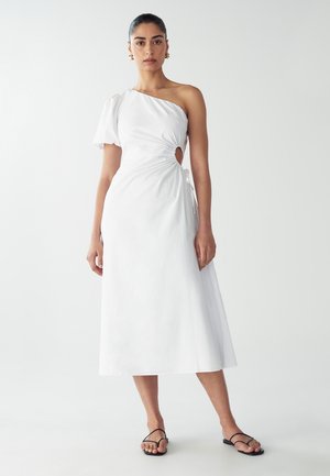 Vestido blanco de un solo hombro con un detalle retorcido y un corte en forma de llave. Tejido suave, diseño hasta el tobillo, acompañado de chanclas negras.