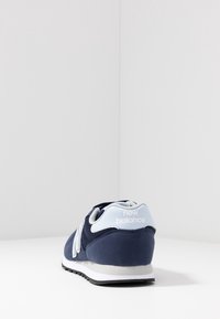 Marinblå atletisk sko med en slät ovandel av tyg, grå accenter, rund tå och en vit sula. Har varumärkeslogotyp på hälen.