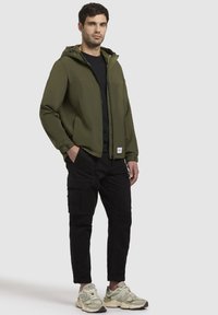 Grüne Kapuzenjacke mit Reißverschluss, schwarzes T-Shirt, Cargo-Hose und hellgrüne Sneakers. Die Jacke hat eine leicht glänzende Oberfläche.