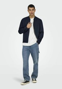 Marineblå bomberjakke med glidelås, kombinert med en hvit t-skjorte, lyseblå jeans og svarte joggesko med hvite såler. Modell stående.