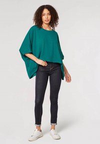 Grünes, oversized Oberteil mit lockerer Passform, kurzen Ärmeln und seitlichen Schlitzen, kombiniert mit dunklen Skinny-Jeans und weißen Sneakern.