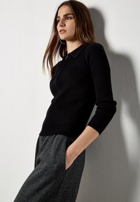 Polo nero a maniche lunghe a coste con colletto, abbinato a pantaloni grigi con disegno. La maglia presenta una chiusura frontale con zip e un design aderente.