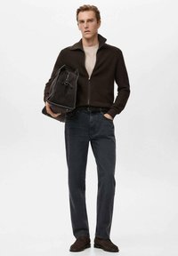 Brązowy teksturowany sweter na suwak, jasna koszula pod spodem, niebieskie dżinsy, ciemne obuwie oraz ciemny plecak materiałowy; neutralne tło.