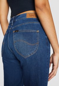 Blå denimjeans med hög midja, som har en brun läderetikett och orange sömmar. Bakfickorna har en distinkt böjd design.
