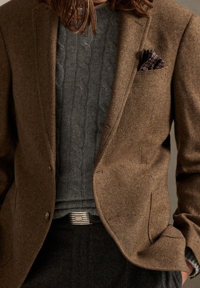 Καφέ σακάκι ψαροκόκκαλο με μαντηλάκι στην τσέπη με σχέδιο, γκρι πουλόβερ πλεκτό με σχέδιο κοτσίδας και σκουρόχρωμο παντελόνι με ορθογώνια αγκράφα στη ζώνη.