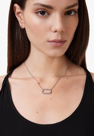 Silver necklace featuring an elongated rectangular pendant with embedded clear stones, resting on a delicate chain. Wearer has a black top.