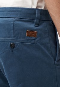 Κοντινή λήψη τσέπης σκούρου μπλε παντελονιού με μαύρο κουμπί και καφέ λογότυπο Timberland στη ζώνη, φορεμένο με άσπρο πουκάμισο.