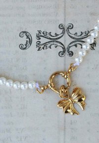 Collana di perle con perle bianche rotonde e un charm a forma di farfalla in oro con un fermaglio circolare, posizionata su uno sfondo testurizzato.