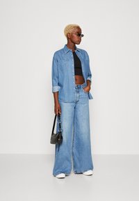 Denim shirt, lichtblauw, lange mouwen, twee borstzakken; zwarte crop top, wijde jeans; zwarte handtas; ronde zonnebril; witte sneakers.