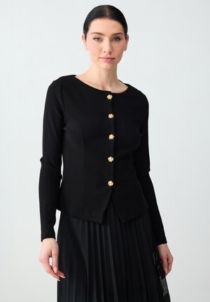 Kobieta z ciemnymi włosami, ubrana w dopasowaną czarną kurtkę z złotymi guzikami i plisowaną czarną spódnicę na jasnym tle.