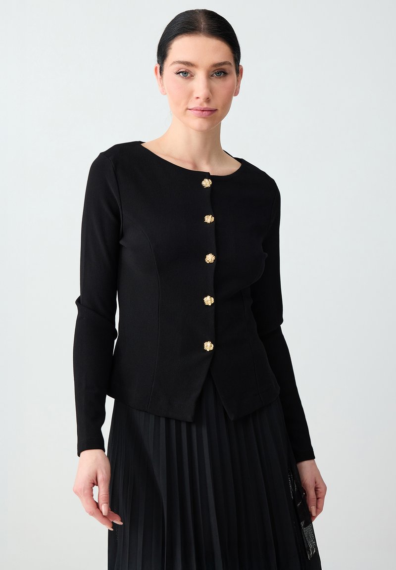 Femme aux cheveux foncés portant une veste noire ajustée avec des boutons dorés et une jupe noire plissée sur un fond clair.