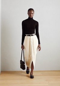 Zwart langemouw turtleneck, crèmekleurige lange rok met een split aan de voorkant, zwarte riem, zwarte handtas en zwarte instappers op een houten vloer.