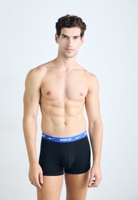 Schwarze Boxershorts mit blauer elastischer Taille und "NIKE"-Logo. Glattes Material, enganliegende Passform und kurzes Bein-Design. Männlicher Model posiert.