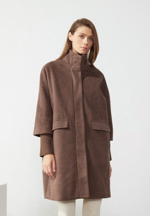 Manteau en mélange de laine marron avec un col haut, des poches latérales et des manches courtes. Texture douce, coupe ample et design minimaliste.