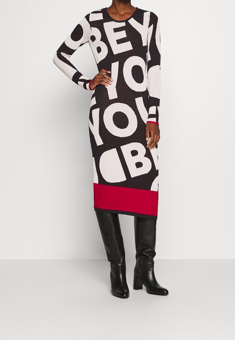 Femme portant une robe midi manches longues noire, blanche et rouge avec de grandes lettres en bloc et des bottes noires hautes jusqu'aux genoux, debout sur un fond blanc.