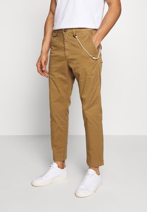 Mann trägt beige Chinos mit einem Schlüsselanhänger aus Seil und weißen Turnschuhen, steht vor einem schlichten hellen Hintergrund, eine Hand in der Tasche.