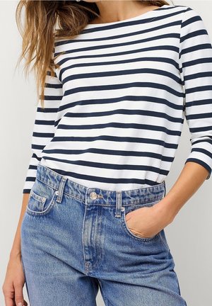 Donna che indossa una maglietta a righe bianche e blu con maniche lunghe, infilata in un paio di jeans blu a vita alta, con una mano nella tasca.