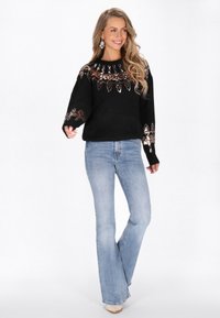 Maglione nero lavorato a maglia con colletto e maniche decorate, abbinato a jeans svasati di un leggero blu. L'outfit presenta paillettes decorative.