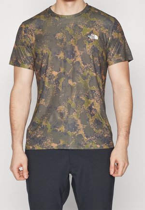 Homme portant un t-shirt à manches courtes à motif camouflage The North Face et un pantalon noir, debout, les bras détendus le long du corps.