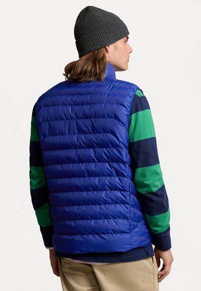 Blauer Puffer-Weste mit gestepptem Muster, getragen über einem grün-blauen gestreiften Langarmshirt. Graue Strickmütze. Lässige Passform.