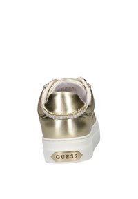 Gouden metallic sneaker met strasaccenten, witte rubberen zool en gestructureerde achterkant; heeft "GUESS" branding op de hiel.