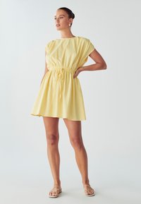 Geltona kelių ilgių suknelė, pagaminta iš lengvos medžiagos, su raišteliu juosmens srityje ir rankovėmis iki pečių, laisvo kirpimo ir glotnios tekstūros.