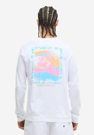 Mężczyzna z zaplecionymi włosami, ubrany w białą koszulę z długim rękawem z pastelową grafiką surfera i napisem "Ocean Pacific" na plecach.