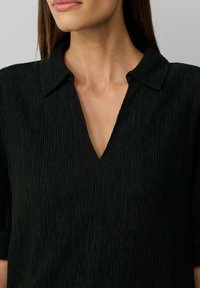 Blouse noire texturée avec un col et une encolure en forme de V. Le tissu présente des lignes souples et froissées ainsi que des manches courtes.