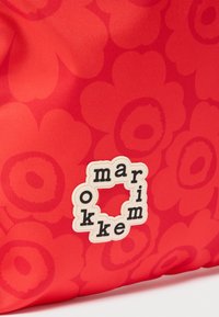 Κόκκινο ύφασμα με floral σχέδιο και λευκή ετικέτα με το λογότυπο που αναγράφει τη λέξη "marimekko" σε σχήμα τετραγώνου.