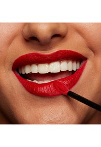 Ljus röd läppstift applicerat på släta läppar, med en svart applikatorborste som rör vid läppen. Vita tänder synliga i ett lätt leende.