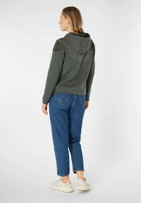 Groene hoodie met een textuurpaneel op de schouders, gecombineerd met blauwe denim jeans en witte sneakers. De achterkant laat het ontwerp duidelijk zien.