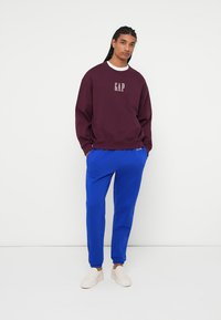 Bordová mikina s logom "GAP", biely okrúhly výstrih, skombinovaná s modrými teplákmi. Model má na sebe béžové slip-on topánky. Hladká tkanina a uvoľnený strih.