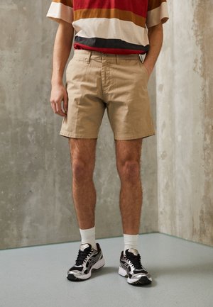 Beżowe szorty o luźnym kroju, wykonane z bawełny, z gładkim przodem. Noszone z pasiastą koszulą w czerwono-brązowe paski oraz czarnymi trampkami i białymi skarpetkami.