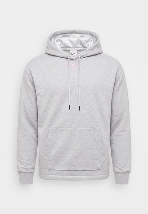 Mężczyzna z blond włosami, ubraną w jasnoszary kaptur z różowym logo i białe spodnie, stojący na jednolitym jasnym tle.