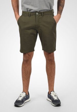 Shorts verde oliva con cierre de botón, dos bolsillos laterales y un ajuste casual. Combinados con zapatillas gris oscuro con cordones blancos.