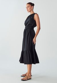 Black sleeveless dress with a gathered neckline, an elastic waistband, and a ruffled hem. Paired with black strappy sandals on bare feet.