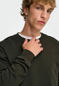 Jeune homme aux cheveux clairs et aux yeux bleus ajustant le col d'un sweat-shirt vert foncé par-dessus une chemise blanche sur un fond uni.
