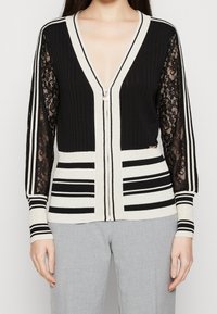 Vrouw draagt een zwart-wit gestreepte ritssluiting cardigan met kanten mouwen, gecombineerd met lichtgrijze broek, getoond van nek tot heupen.