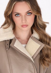 Giacca in pelle color tan con collo e foderato in shearling crema, caratterizzata da una chiusura a zip e dettagli con bottoni a pressione, messa in risalto da una morbida texture.