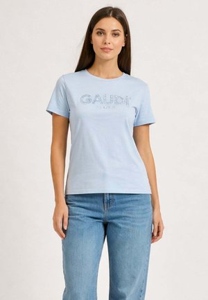 Donna che indossa una maglietta a maniche corte di colore azzurro chiaro con la scritta "GAUDI" e jeans blu medio su uno sfondo chiaro e semplice.