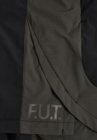 Mørkegrønt og sort stof med ternet mønster, synlig søm og "F.U.T." trykt i gråt på den grønne sektion.