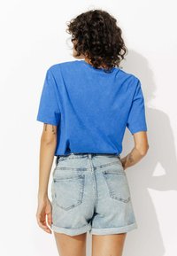 Personne aux cheveux bouclés portant un t-shirt bleu et un short en denim clair roulé, se tenant de dos face à la caméra devant un fond blanc.