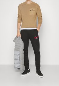 Beige Sweater mit Brusttasche, schwarze Jogginghose mit rotem Logo, graue Steppjacke und schwarze Sneakers mit weißen Sohlen.