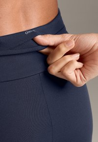 Colanți de yoga elastici, de culoare bleumarin, cu un brâu pliabil și cusături discrete. O mână este văzută ajustând brâul pentru un fit perfect.