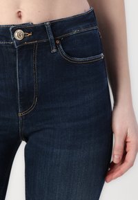 Dark denim jeans with a mid-rise waist, featuring orange stitching, front pockets, and a metal button closure. Smooth texture and fitted shape.