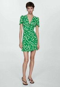 Rochie florală verde cu mâneci scurte, decolteu în V, talie plisată, tiv evazat și margine volanată. Asortată cu flip-flops negri.